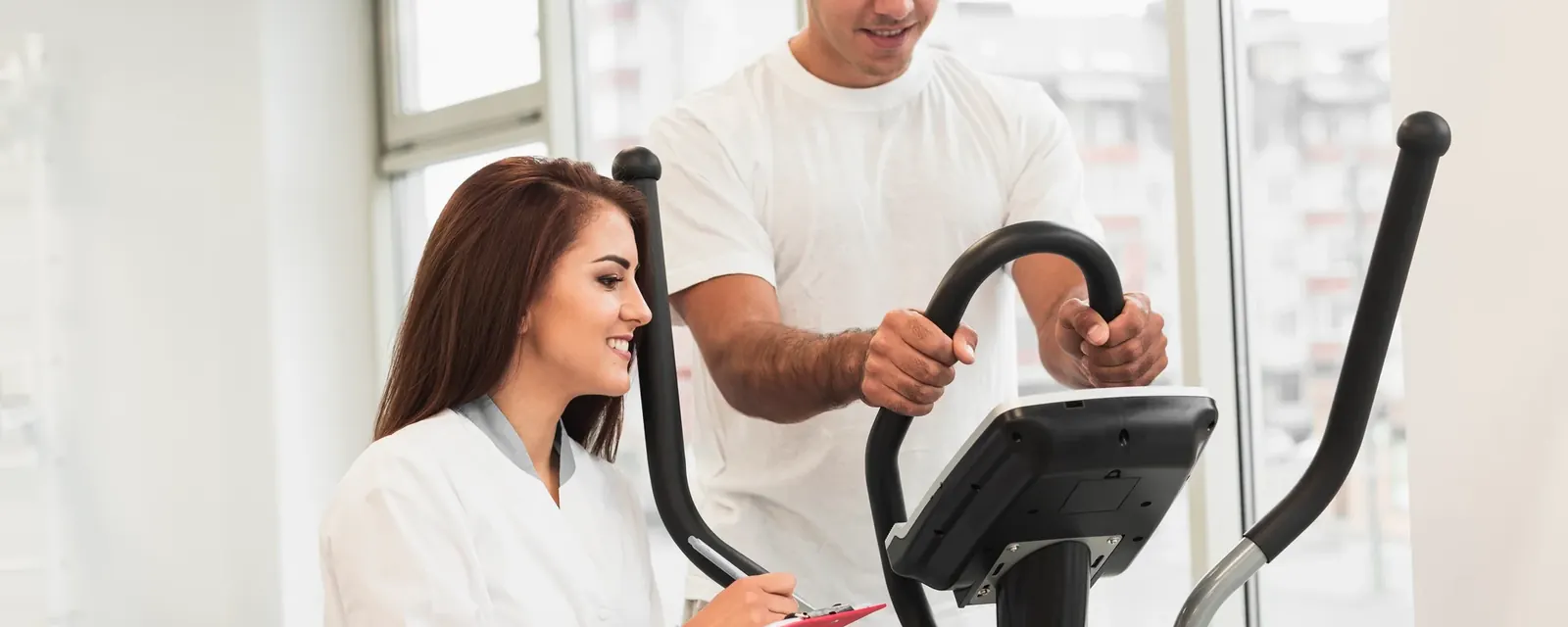 An exercise physiologist recording measurements while a patient uses a cross-trainer